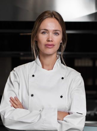 A woman in a chef 's uniform is standing in a kitchen with her arms crossed.