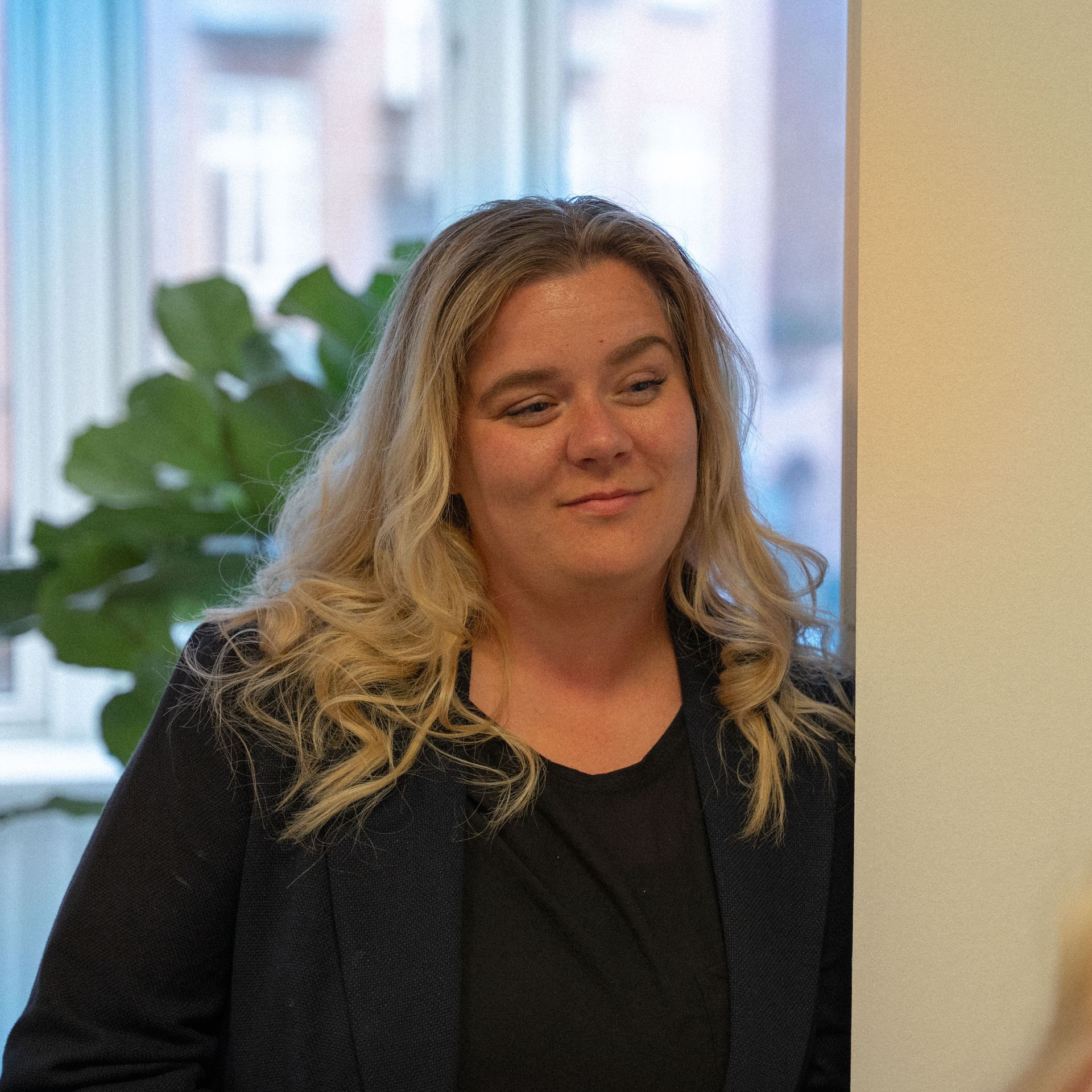 Een vrouw in een zwart jasje leunt tegen een muur en glimlacht.