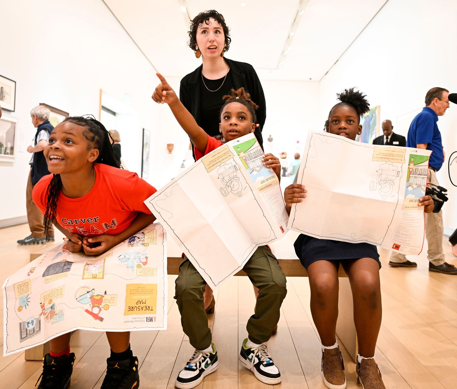 A museum guide points toward art for two children sitting on a bench, both holding illustrated activity maps.