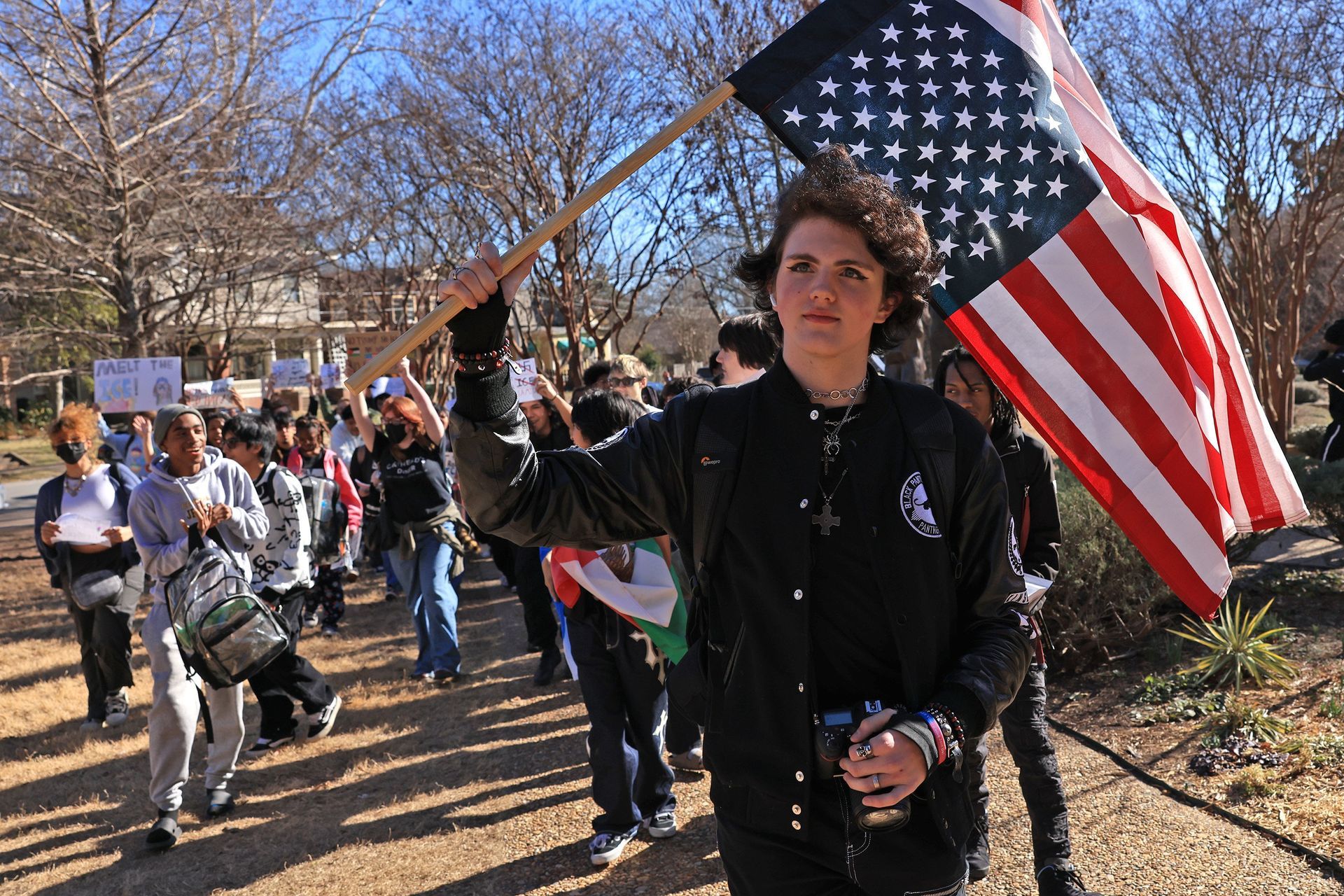 A person walking outdoors holding an American flag with a group of people marching in the background.