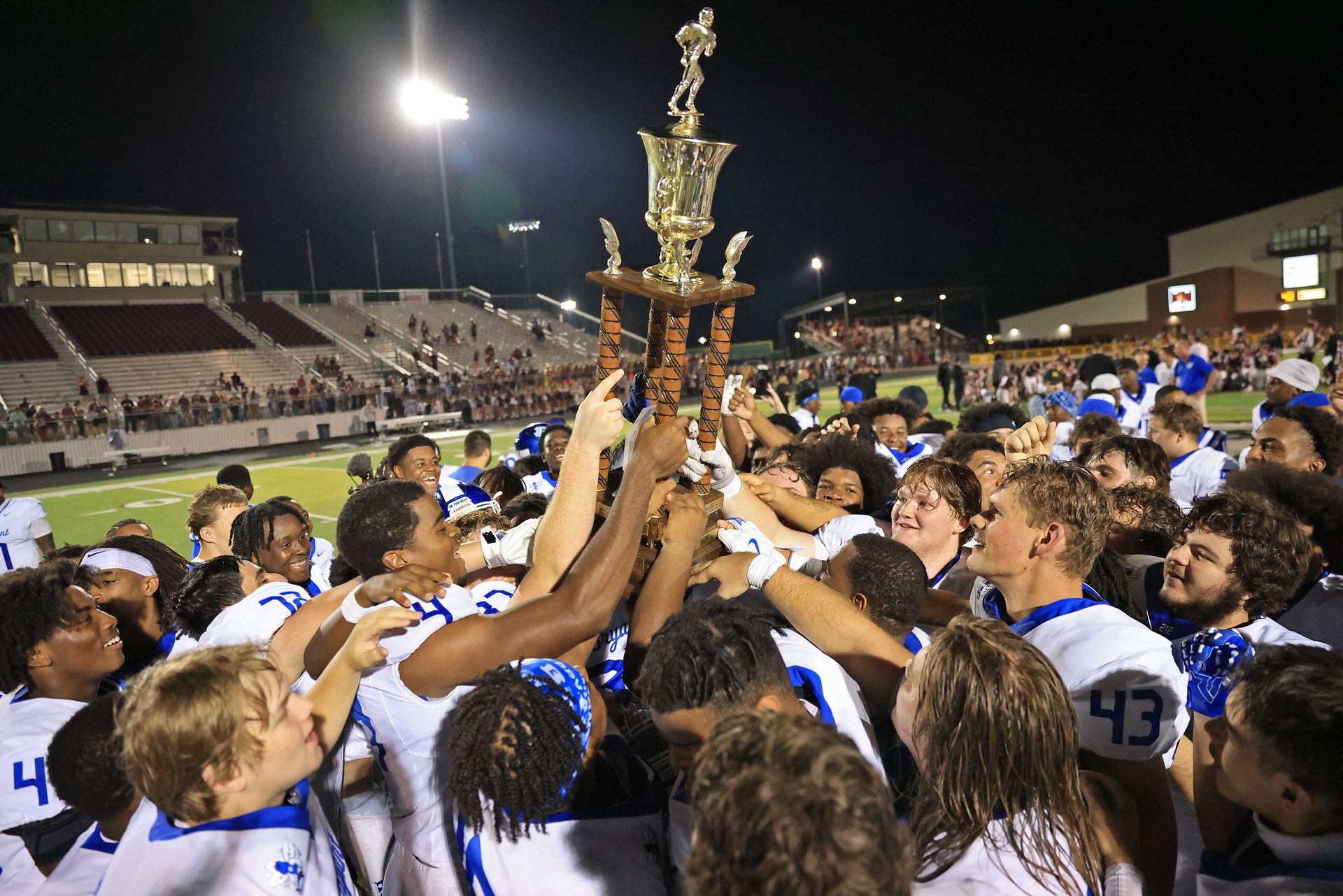 A football team in white jerseys celebrates a victory at night, lifting a large gold trophy in the center of the field.