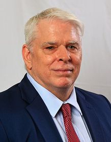 A man in a blue suit, light blue dress shirt, and red patterned tie, posed against a plain, neutral background.
