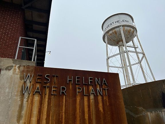 The West Helena Water Plant sign on a rust-streaked wall, with a white water tower standing against a gray, cloudy sky.
