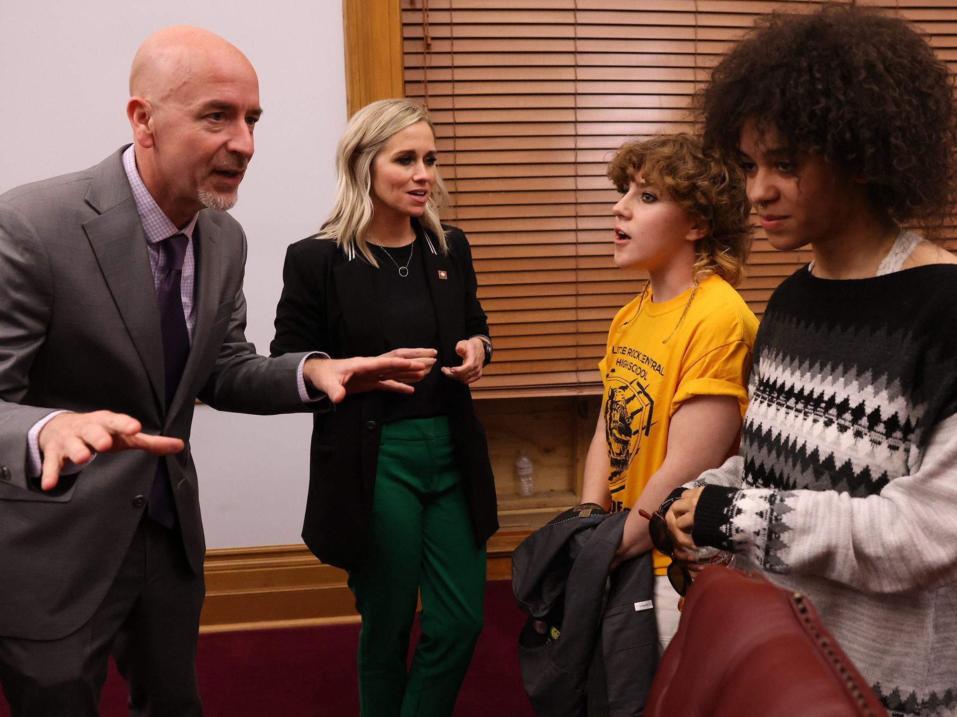 A man in a suit speaks to a woman and two individuals in an office setting with wood-slatted blinds in the background.