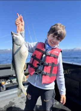Boy in life vest holding up a large fish on a boat, smiling, with blue sky and water in the background.