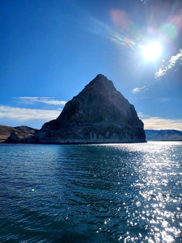 Pyramidal rock formation rises from blue water under a bright sun.