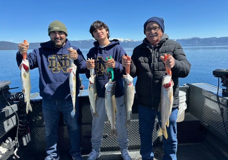 Three people on a boat holding fish they caught, blue water and mountains in background.