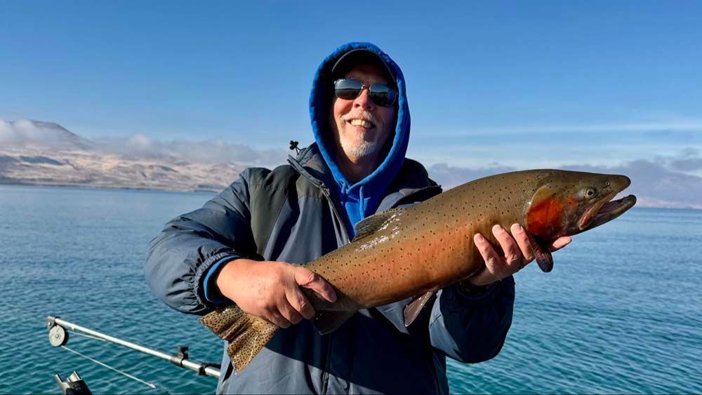 Man in blue hoodie holding a large fish, with lake and mountains in the background.
