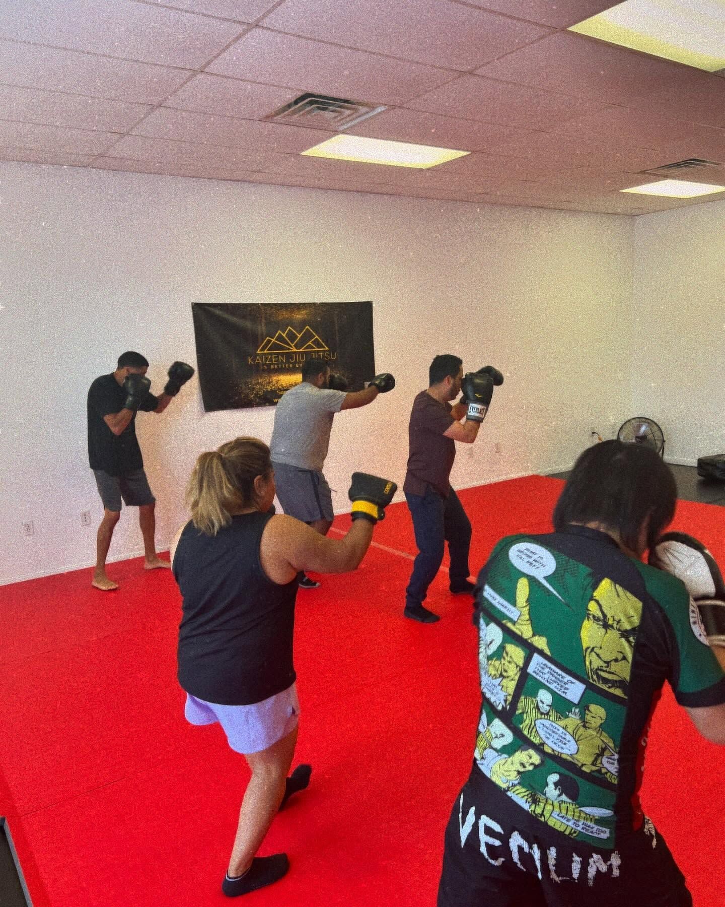 People practicing boxing drills in a red-floored training room, wearing gloves and facing a wall target.