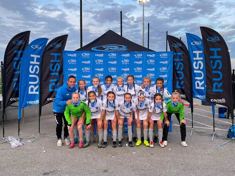 Soccer team in white and blue uniforms with medals posing in front of a banner.