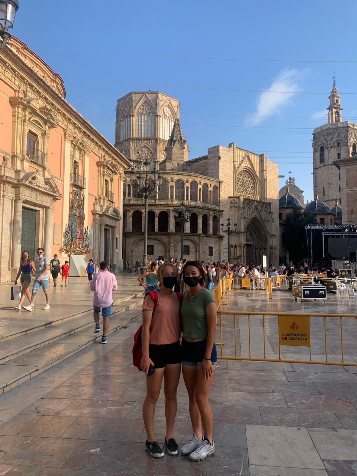 Two people posing in a plaza with a cathedral in Valencia, Spain. They wear masks and shorts.