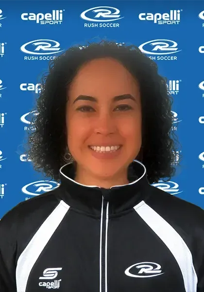 Woman with curly hair, smiling, wearing a black and white athletic jacket. Background has Rush Soccer logos.