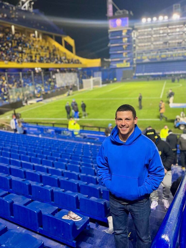 Man in blue hoodie smiles at soccer stadium with blue and yellow seating.