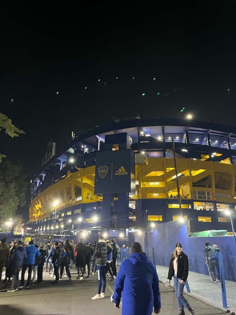 Night view of La Bombonera stadium in Buenos Aires, Argentina, with fans walking outside, lit in yellow and blue.