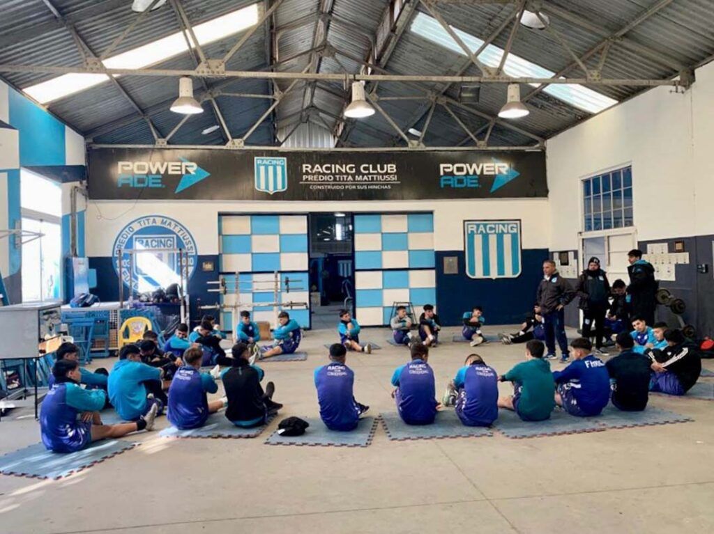 Soccer players in blue and light blue uniforms seated on mats in a gym.