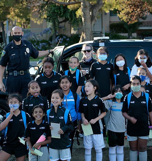 Officer with group of children posing near a police car. Children wear soccer gear and masks.