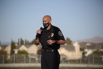 Police officer in uniform speaking into a microphone outdoors, wearing a mask.