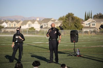 Two police officers speak on a football field; a speaker is nearby. Houses and a hill are in the background.