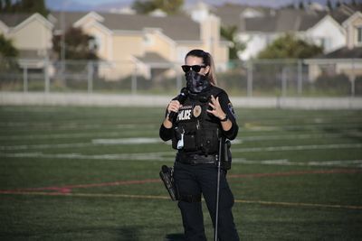 Police officer in tactical vest, on a green field, wearing face covering and sunglasses. Houses in background.