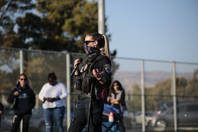 Woman in uniform speaks into microphone outdoors; people in background.
