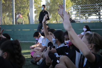 Children seated on grass with arms raised at a sports field, some wearing masks.