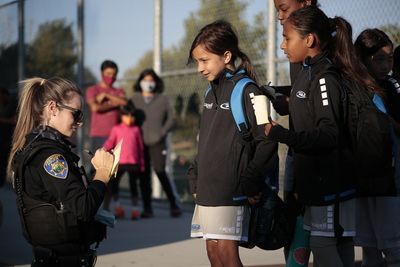 Police officer writing on a notepad, talking to children near a soccer field. Spectators in background.