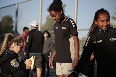 A police officer writing on a notepad next to a girl in a soccer uniform; other people in the background.
