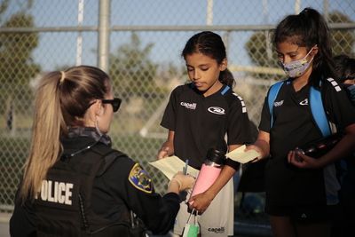 Police officer handing paperwork to two soccer players outdoors near a fence.