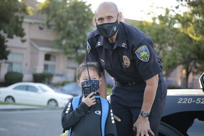A police officer in mask smiles beside a child with a radio, near a parked car.