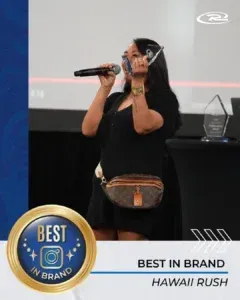 Woman with microphone, holding award, wearing a black dress and fanny pack. 