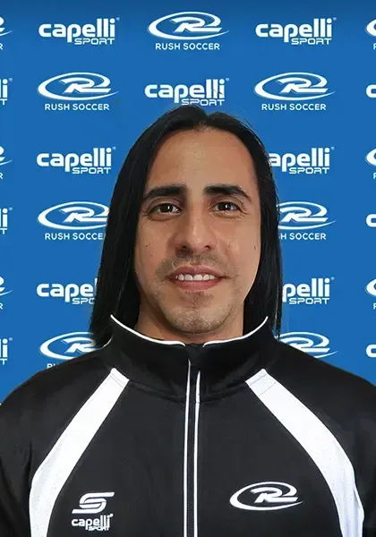 Man with long dark hair wearing a black and white track jacket, posing in front of a blue and white logo background.