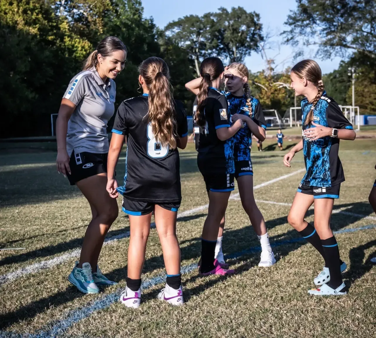 Soccer team huddles with coach on green field. Players wear blue/black jerseys, coach in grey. Smiling, laughing.