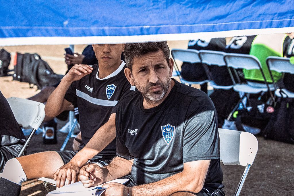 Soccer coach in black shirt and assistant under a canopy, reviewing notes during a game.