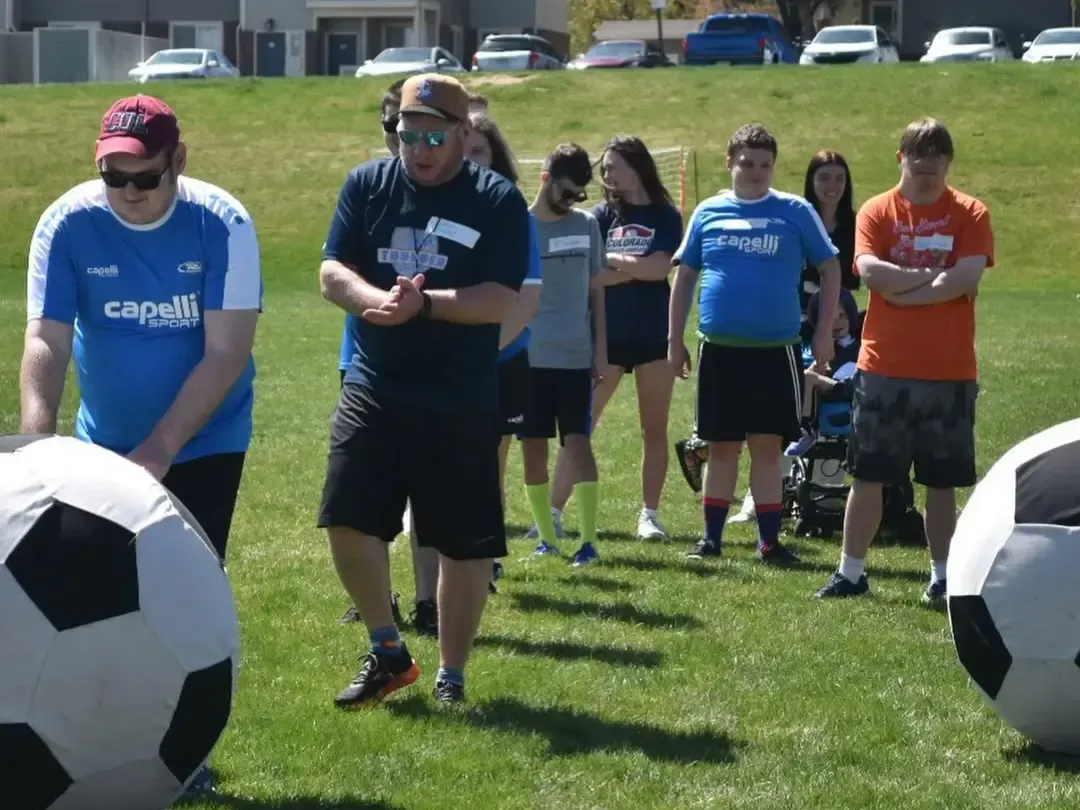 People on a grassy field with large soccer balls. Man in hat gestures. Others watch, some in athletic attire.