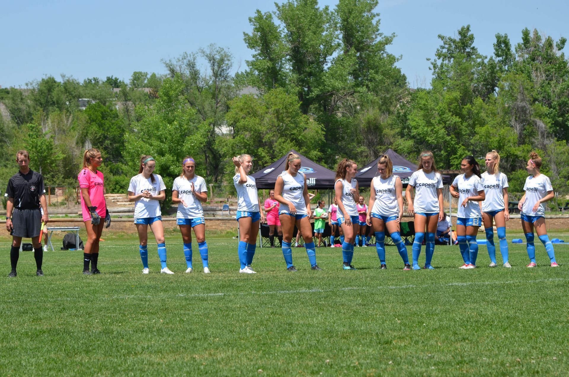 Soccer players in blue and white uniforms on a grassy field, with referees and tents in the background.