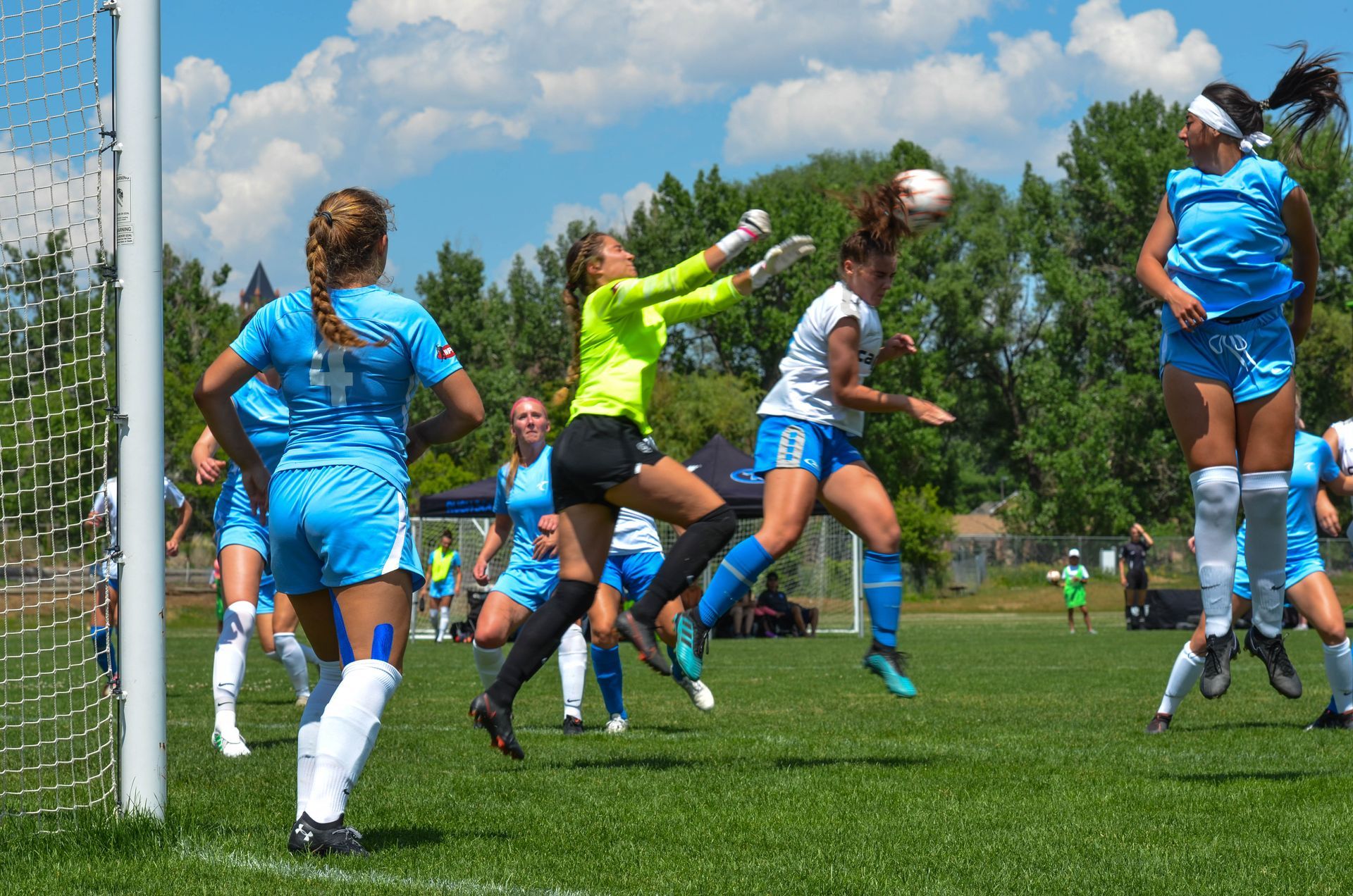 Soccer players in blue and white uniforms jump for a header near the goal; goalie in neon green.