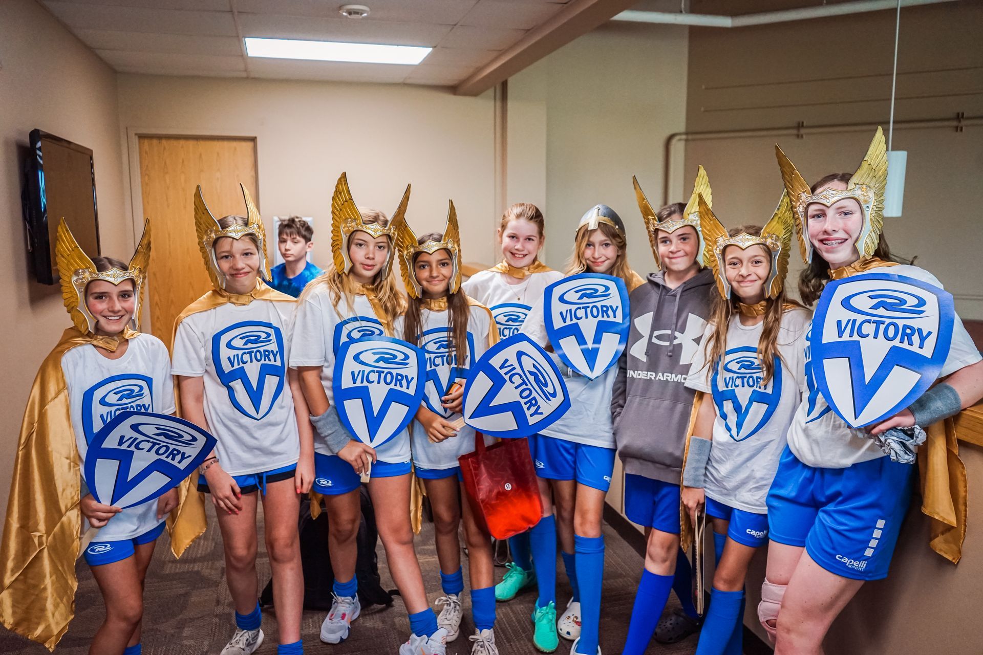 Group of children in matching outfits posing, wearing superhero helmets, holding shields.