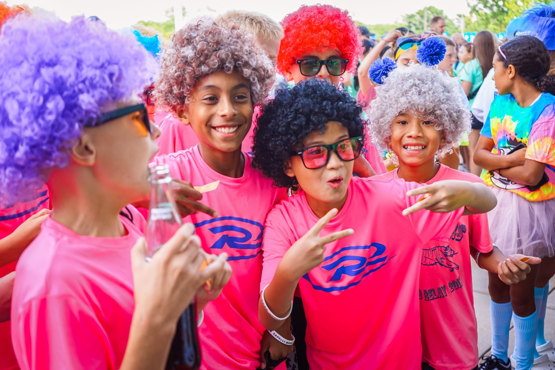 Group of children in pink shirts, colorful wigs, and sunglasses posing outside.