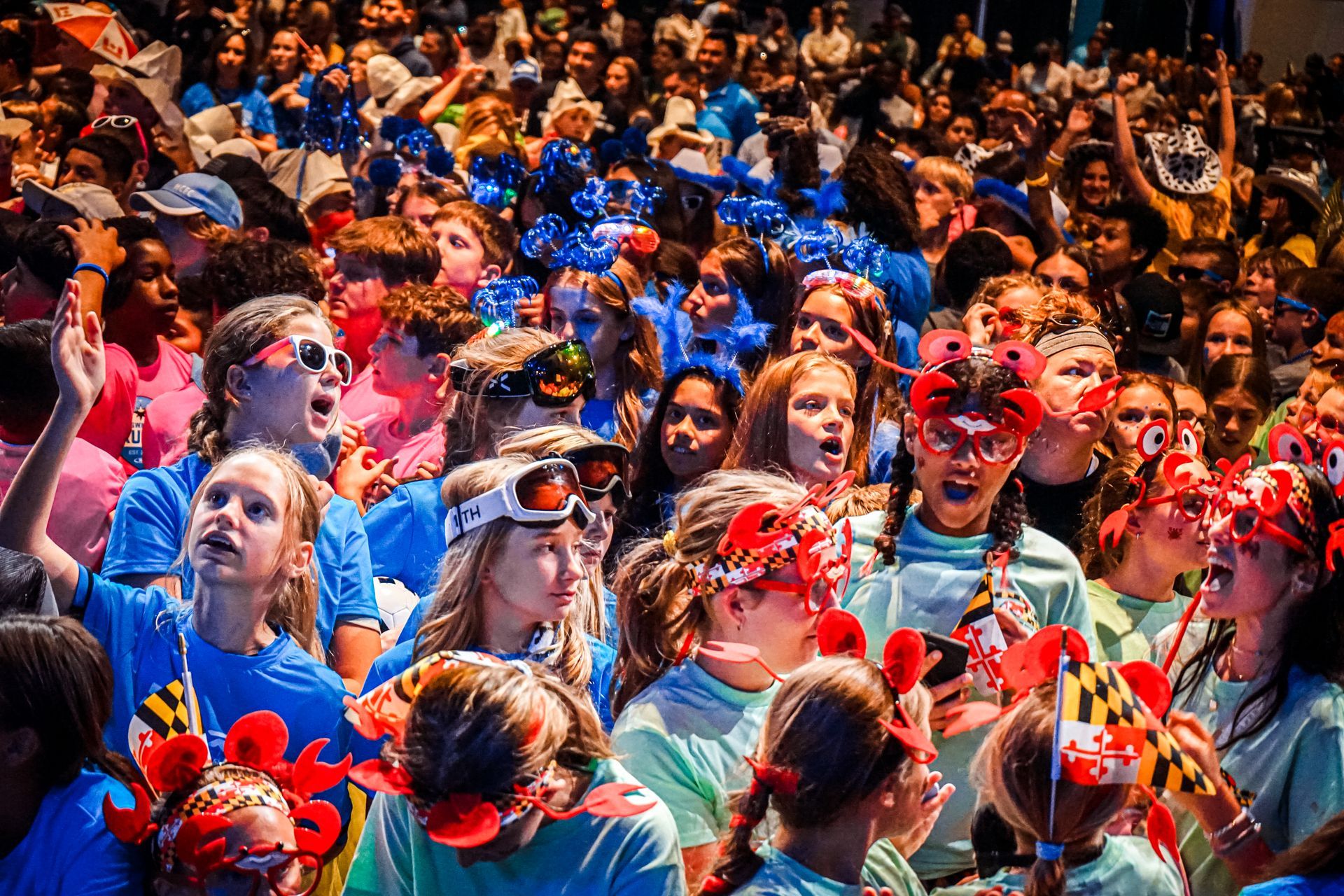 Large crowd of people wearing blue and green shirts, some with crab-themed headbands and goggles, cheering enthusiastically.