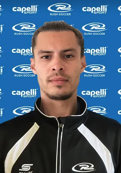 Man with pulled-back hair wearing a black and white track jacket, in front of a blue background with soccer logo.