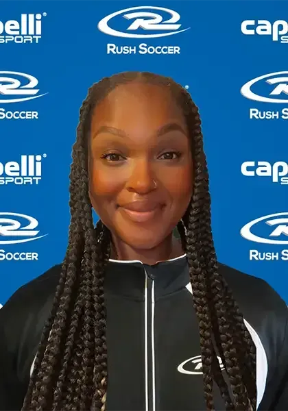 Woman with long braids smiles, wearing a black and white jacket, standing in front of a blue backdrop with soccer logos.