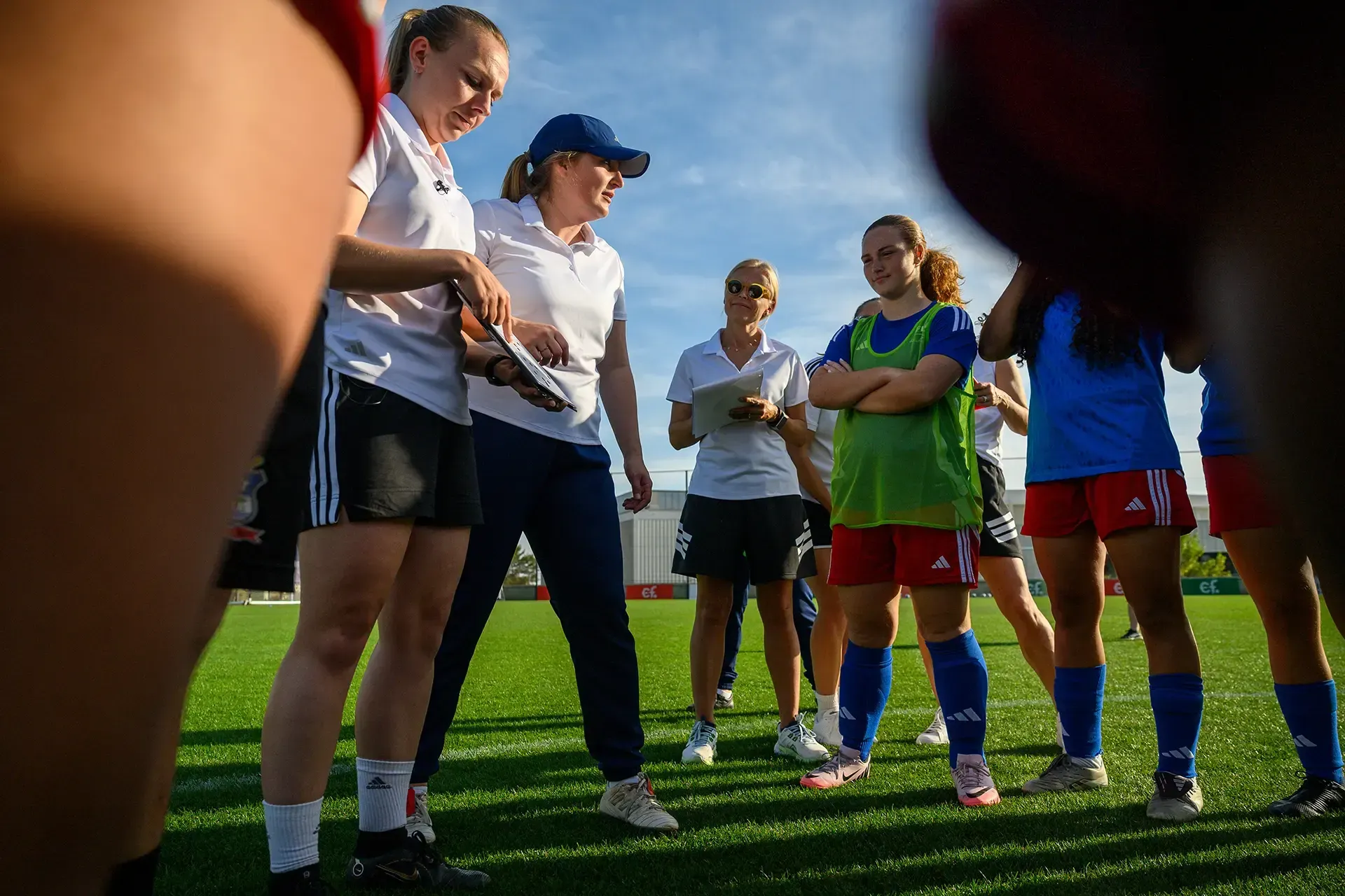 A soccer coach talking with players on a green field. Players listen, some with arms crossed.