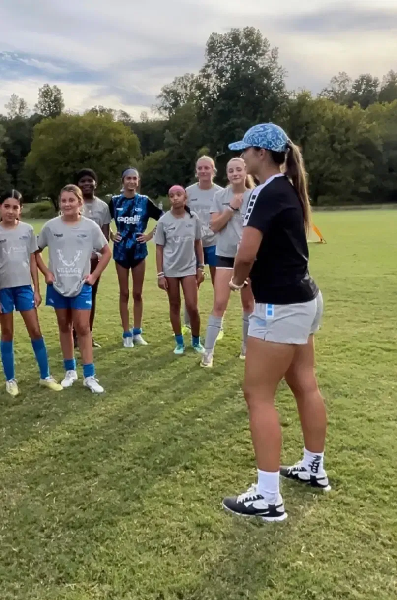Soccer coach addressing a team on a grassy field. Players wear gray and blue attire. Coach in black and gray.