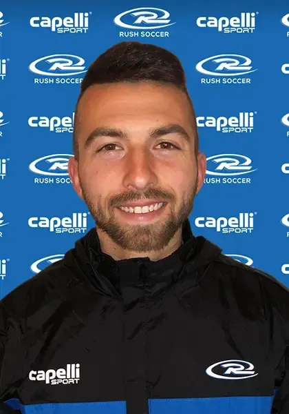 Man with dark hair and a beard, smiling, wearing a jacket, in front of a blue banner.