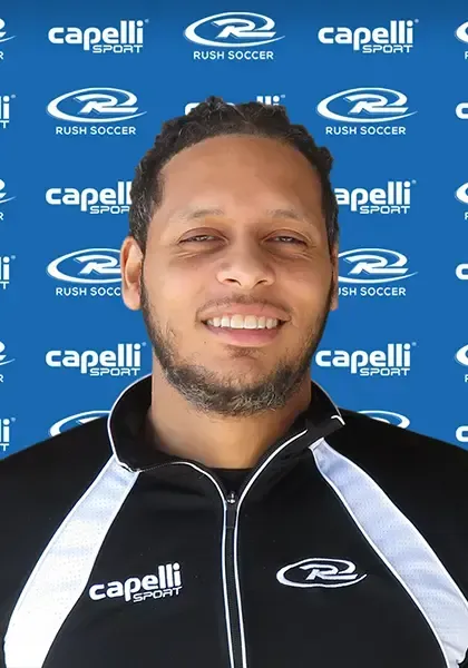 Man in black and white jacket smiles in front of a blue and white background with the Rush Soccer logo.