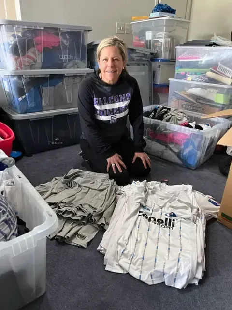 Woman kneeling amidst boxes and piles of clothes; smiles.