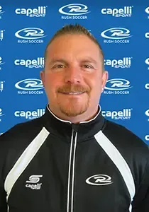 Man in a black and white jacket, smiling, in front of a blue background with soccer logos.