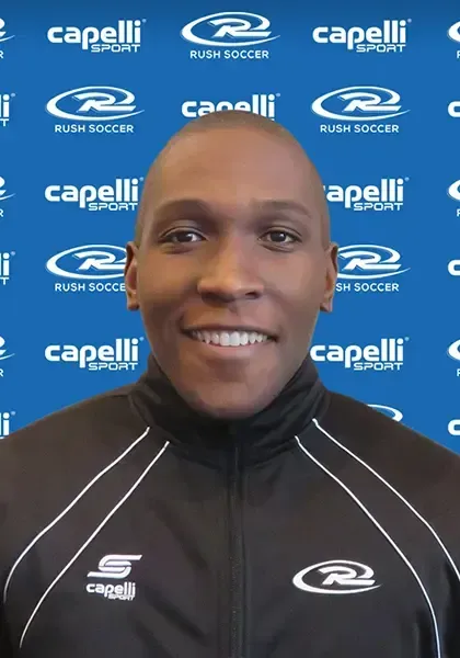 A person wearing a black jacket with a white collar, smiling. Background features 