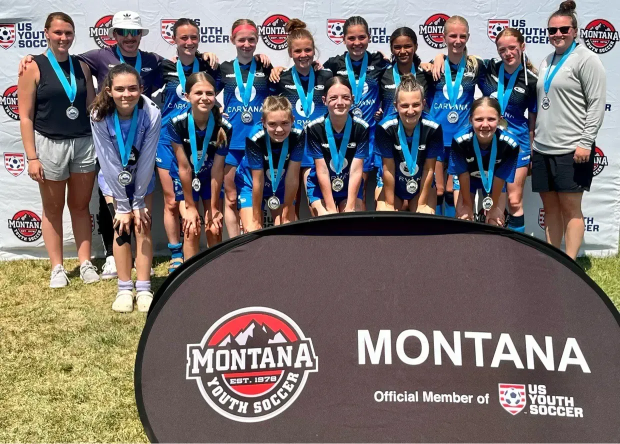 A youth soccer team in blue and black uniforms poses with medals in Montana, in front of a banner.