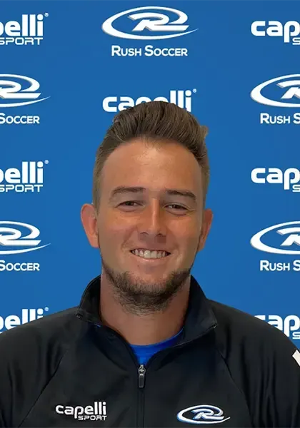 A man in a soccer jacket smiles in front of a blue backdrop with soccer-related logos.